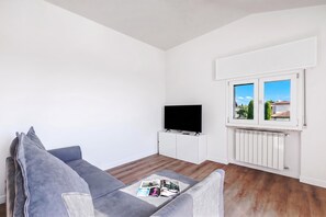 A living room with a grey sofa, a flat-screen TV on a white cabinet, and a window with a view of a house and trees. - Matta Apartment 400m From The Lake (Sirmione)