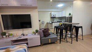 Un saló modern amb televisió plana, un sofà, una zona de menjador amb taula i tamborets, i una cuina al fons.