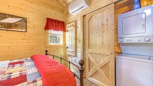 Quarto de cabana de madeira com uma cama, ar condicionado, máquina de lavar e uma janela com cortina vermelha.