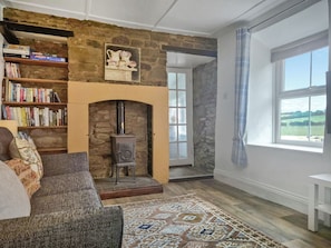Ein gemütliches Wohnzimmer mit Kamin aus Stein, Bücherregalen, einer Couch und einem großen Fenster mit Blick ins Freie.