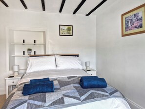 Una cama bien hecha con una colcha de estampado geométrico y dos toallas dobladas. Hay dos mesitas de noche con lámparas y un estante pequeño con una planta.