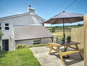 Una casa con techo de tejas, un pequeño jardín y una mesa de madera con sombrilla.