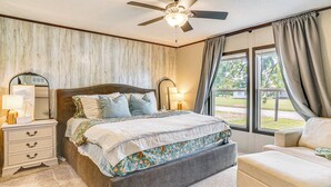 Une chambre à coucher comprenant un lit, des tables de chevet, une commode, une chaise, un ventilateur de plafond et de grandes fenêtres avec des rideaux.
