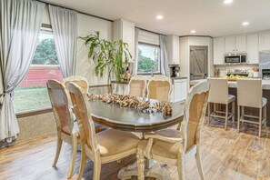 Ein Essbereich mit einem runden Tisch und vier Stühlen, eine Küche mit weißen Schränken und einer Mikrowelle, sowie ein großes Fenster mit Vorhängen.