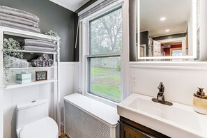A bathroom with a toilet, a window, a shelf with towels, a radiator, and a sink with a faucet. - Near Lake & Wineries: Spacious Union Springs Home! (Union Springs)