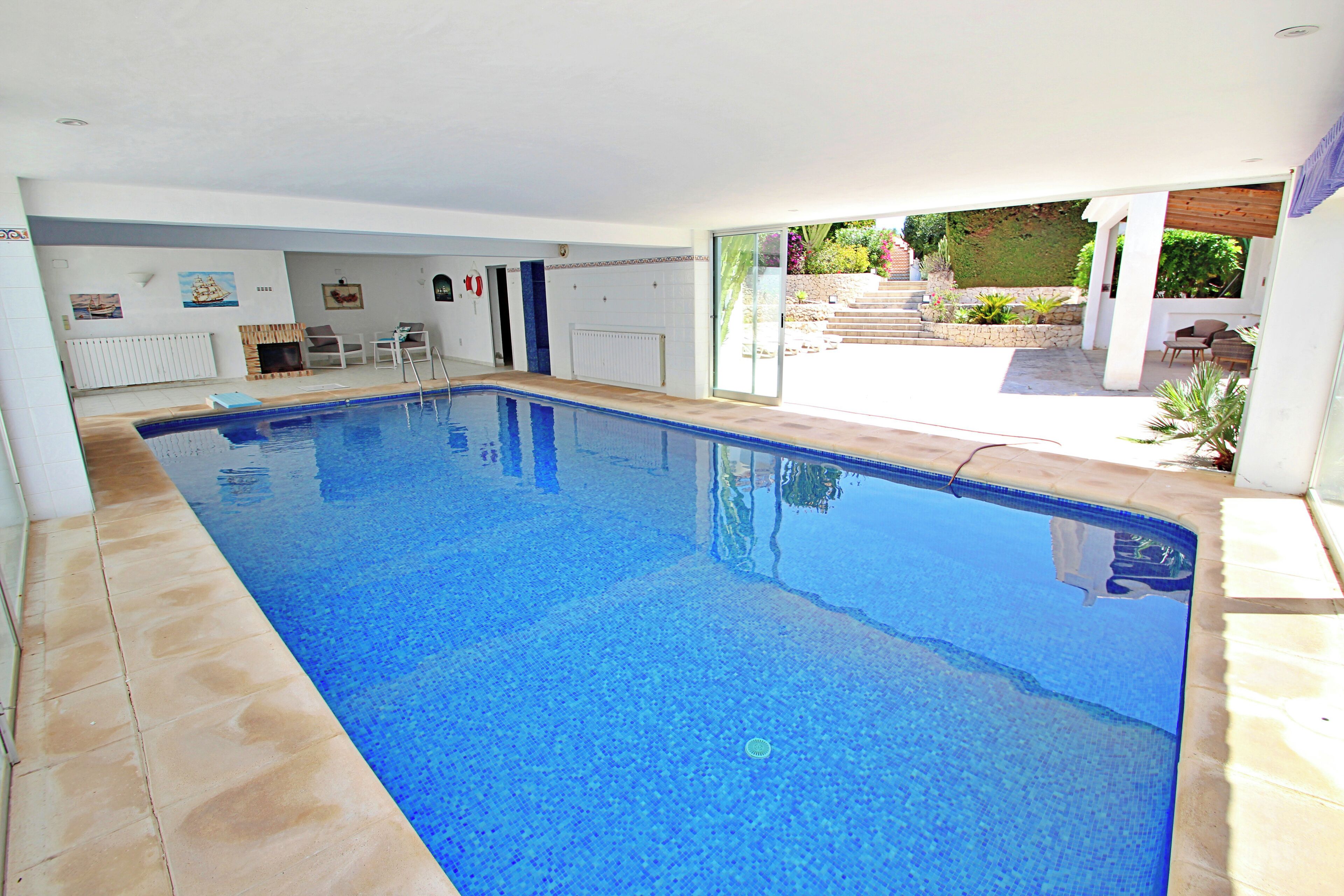 A swimming pool with a tiled deck, a covered patio area, and a view of a garden with steps leading to a door.