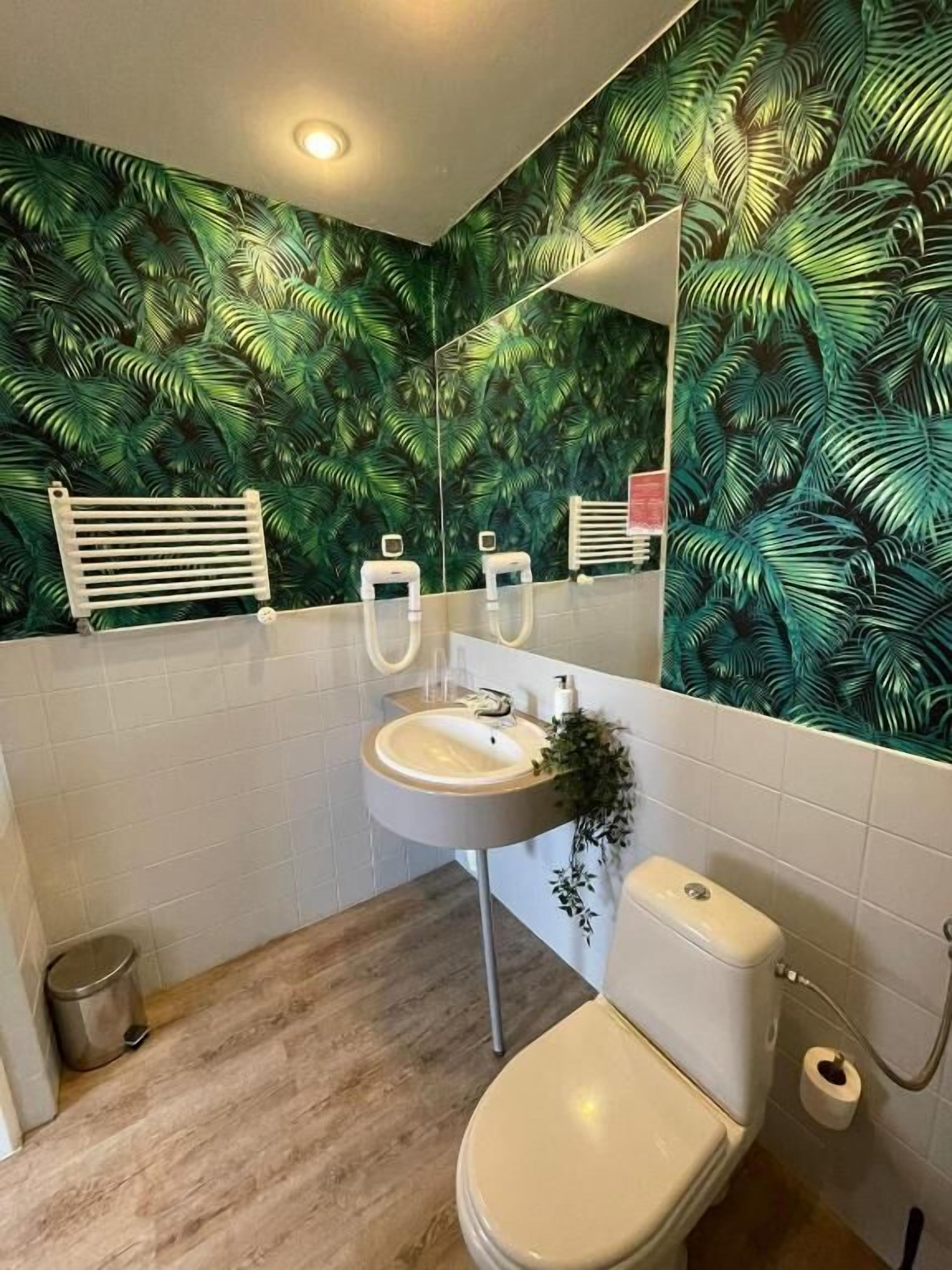 A bathroom with a white sink, toilet, and a mirror above the sink. The walls are covered with a green leafy wallpaper.