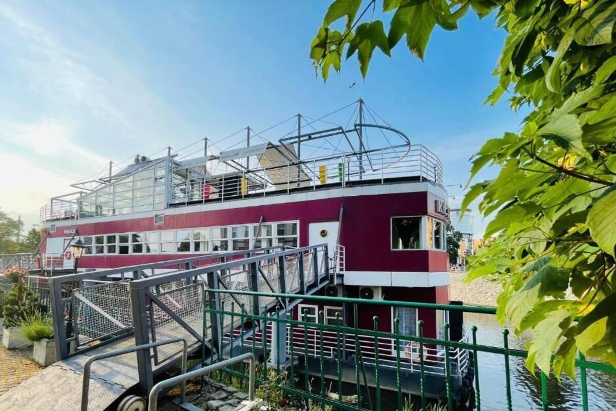 Ein kastanienbraunes und weißes Gebäude mit einer Treppe, die zu einem erhöhten Deck führt, umgeben von Grünflächen und blauem Himmel.