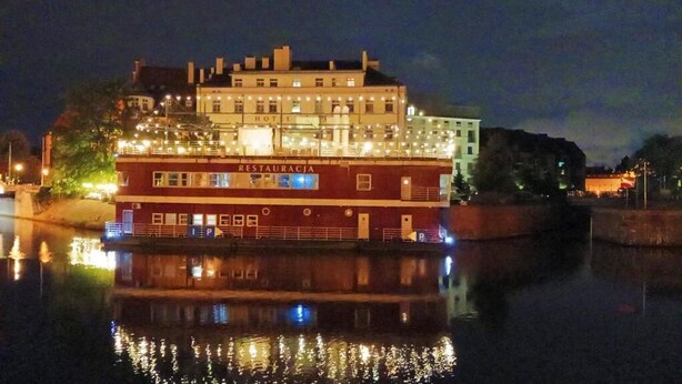 Een groot, meer verdiepingen tellend gebouw met een restaurant op de begane grond, met een opvallend bord en een spiegeling in het water.