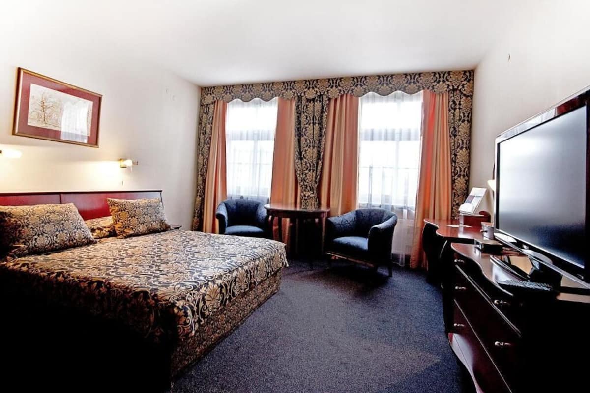 Ein Hotelzimmer mit Bett, zwei Sesseln, einem kleinen Tisch, einem Fernseher und Vorhängen.