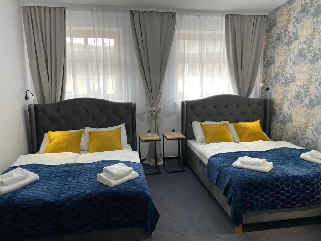 A hotel room with two beds, each with a dark headboard and yellow pillows. There are two bedside tables with lamps, and a small table between the beds.