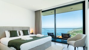 A modern hotel room with a large bed, a seating area with a view of the sea, and a balcony with a table and chairs. - GuestReady - Sunset Heaven Villa (Calheta)