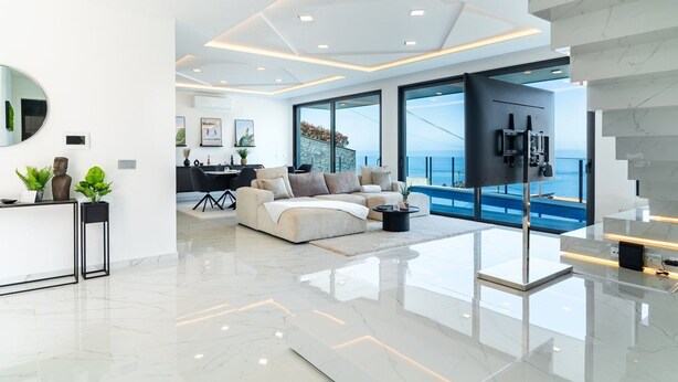 A modern living room with a large sofa, a coffee table, and a TV. There is a dining area with a table and chairs, and a view of the ocean through large windows. - GuestReady - Sunset Heaven Villa (Calheta)