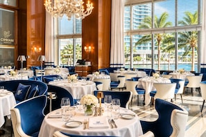 A restaurant with round tables, blue upholstered chairs, and a view of palm trees and buildings through large windows. - LUXURY 1BR/1BA ICON-BRICKELL MIAMI (Miami)