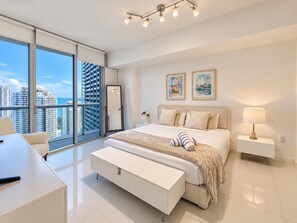A modern bedroom with a large bed, bedside tables, a desk, and a view of high-rise buildings.