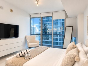 Un dormitorio moderno con una cama grande, un televisor de pantalla plana, una silla y vistas a la ciudad a través de ventanales.