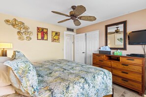 Ein Schlafzimmer mit einem Bett, einer hölzernen Kommode, einem Deckenventilator, einem Fernseher und Kunstwerken an den Wänden.