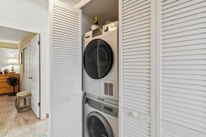A laundry room with a front-loading washer and dryer, a small stool, and a fan.