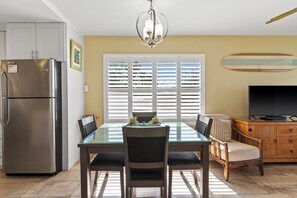 A modern kitchen with a stainless steel refrigerator, a dining table with chairs, a chandelier, and a television on a wooden cabinet.