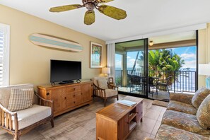 Una sala de estar con televisor, muebles de madera, ventilador de techo y balcón con vistas a palmeras.