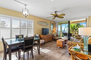 A spacious living area with a dining table, chairs, a sofa, and a television. There is a ceiling fan and a view of the outdoors through large windows. - Kihei Beach #210 – Oceanfront Gem with A/C, Lanai, Pool and Sugar Beach Access (Kihei, Maui)