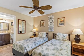 A hotel room with two beds, a ceiling fan, a bathroom with a vanity, and decorative wall art. - Kihei Beach #210 – Oceanfront Gem with A/C, Lanai, Pool and Sugar Beach Access (Kihei, Maui)