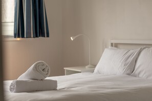 A neatly made bed with white linens and a rolled towel, a bedside table with a lamp, and a window with blue curtains.