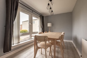 A modern dining area with a wooden table and chairs, large windows with curtains, and a floor lamp. - Fraser Avenue (No 16) (St Andrews)