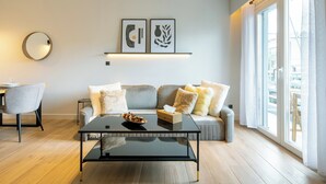 Una sala moderna con una mesa de centro de vidrio, un sofá con cojines, un espejo redondo en la pared y dos obras de arte enmarcadas sobre una repisa.