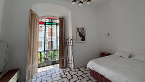 A bedroom with a bed, a nightstand, a framed picture, a unique chandelier, and a view through a window with a balcony.