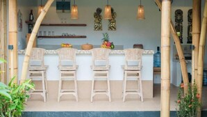 Zona de bar con tres sillas de mimbre, una barra con una exposición de frutas y pilares de bambú.