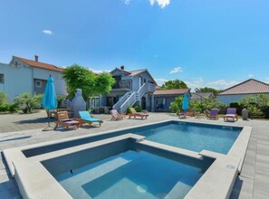 Piscina con sillones de playa y sombrillas, rodeada de casas y un cielo despejado.