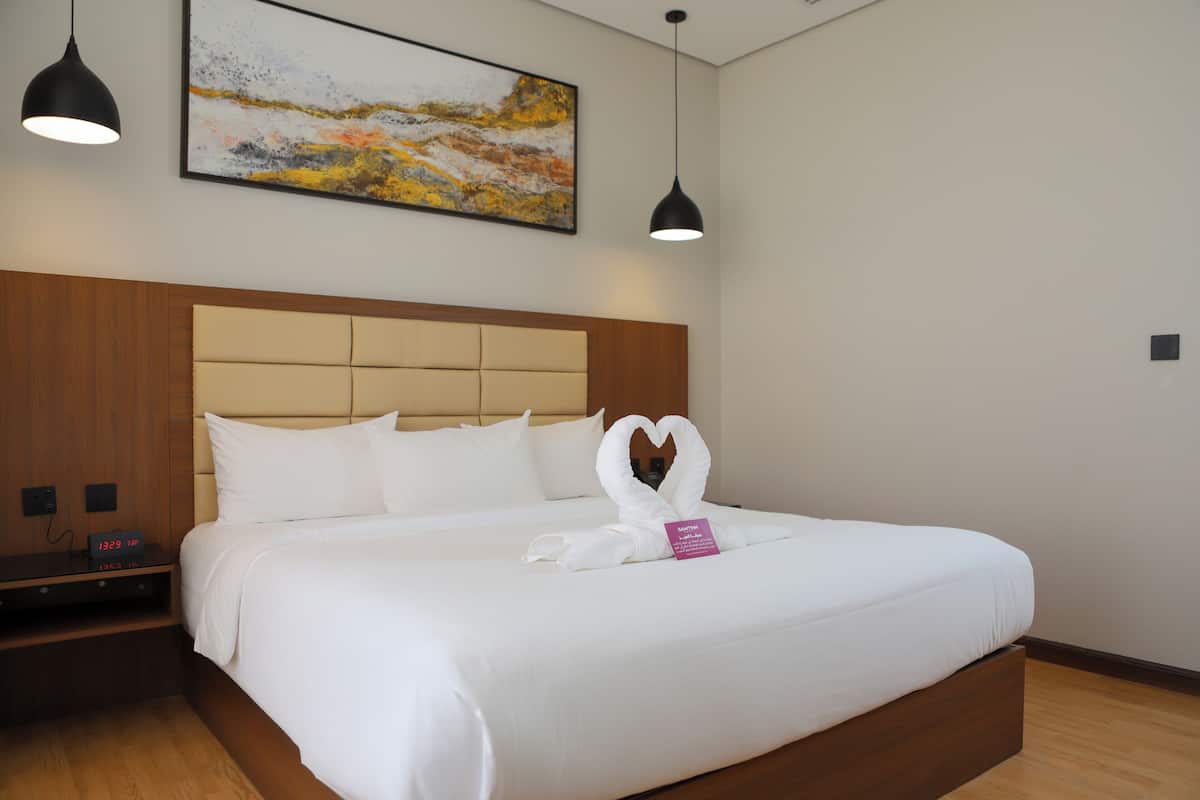 A neatly made bed with a heart-shaped towel arrangement, a wooden headboard, and a painting above.