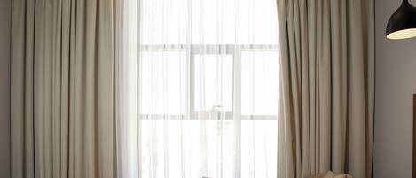 Ein Hotelzimmer mit großem Fenster, einem Bett, einem Sessel und einem kleinen Tisch mit Buch und Schild.