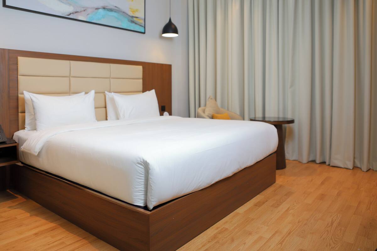 A neatly made bed with a wooden headboard, a bedside table with a lamp, and a view of a curtain and wall art.