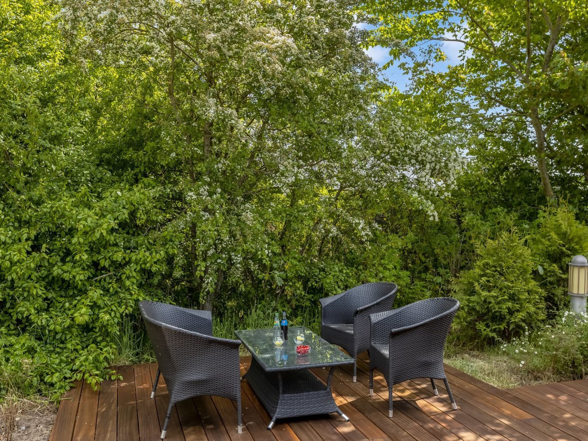 Een terras buiten met een glazen tafel en rieten stoelen, omringd door weelderige begroeiing.