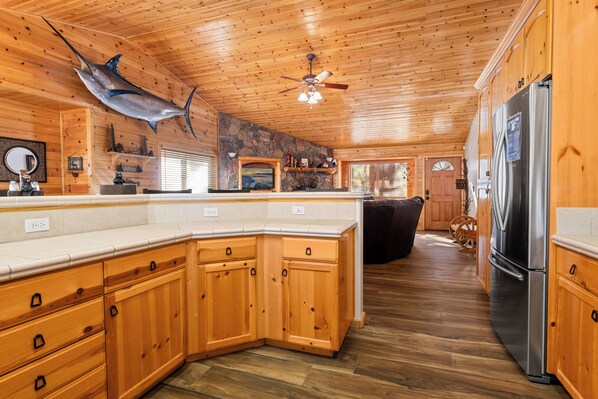 A wooden cabin interior with a large kitchen, stone fireplace, and a ceiling fan.