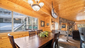 A wooden cabin interior with a dining table, chairs, a chandelier, a stone fireplace, and a kitchen area.