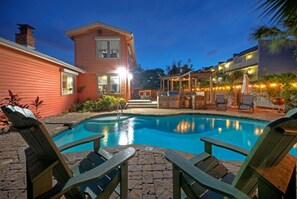 A swimming pool with lounge chairs, a covered patio area, and a two-story building with a visible staircase.