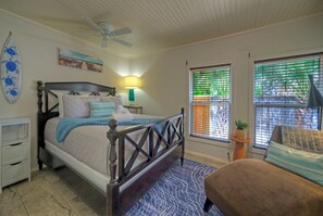 Un dormitorio con cama, ventilador de techo, ventana con persianas, mesita de noche y una pequeña mesa con una planta.
