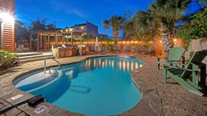A swimming pool with a wooden deck, lounge chairs, and a building with outdoor lighting.