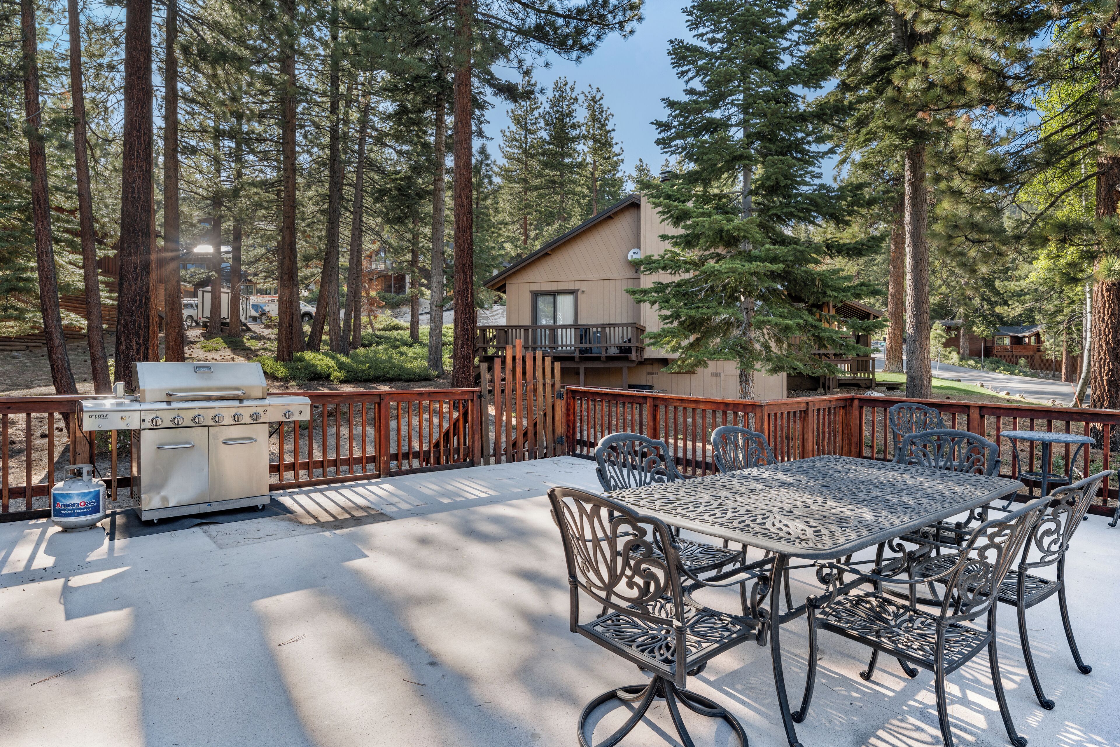 An outdoor patio with a grill, metal table, and chairs, surrounded by tall trees and a wooden fence.