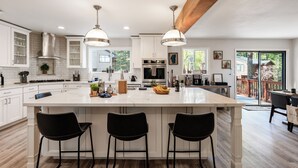 Cocina amplia con una isla grande, muebles blancos y electrodomésticos modernos. Hay tres taburetes negros en la isla y una viga de techo de madera.