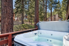 Un jacuzzi en una terraza de madera con vistas a un bosque y una cabaña al fondo.