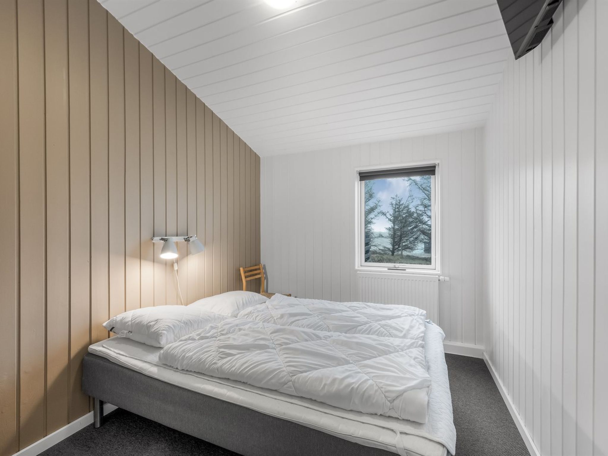 A bedroom with a bed, a nightstand, a chair, a window, and a wall-mounted light fixture.