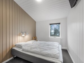 A bedroom with a bed, a nightstand, a chair, a window, and a wall-mounted light fixture.