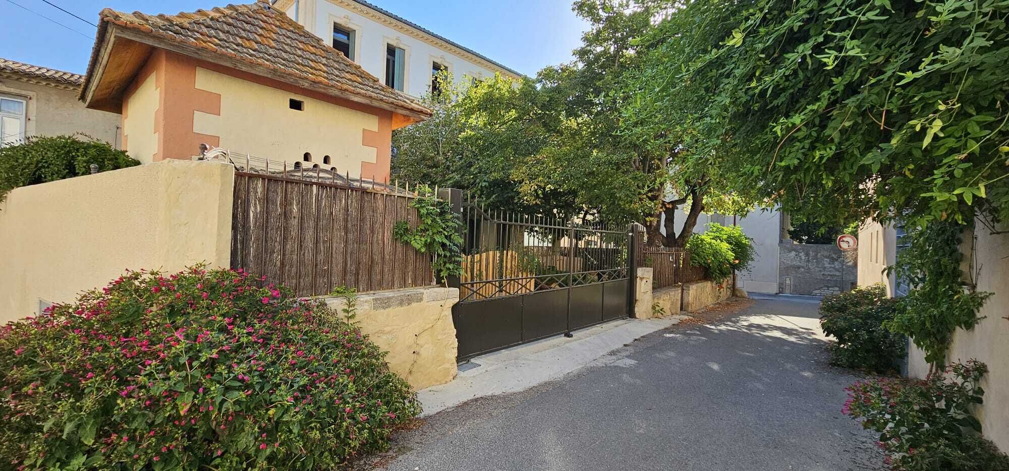 A narrow street with a gated entrance, a building with a tiled roof, and lush greenery.