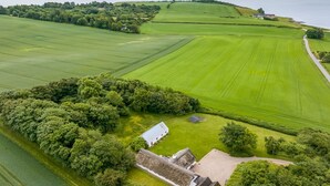 Une vue aérienne d’une propriété rurale, avec de vastes champs verdoyants, un petit bâtiment doté d’un toit de chaume et une étendue d’eau en arrière-plan.