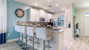 A modern kitchen with white cabinets, a central island with bar stools, a clock on the wall, and a window with a view. - Comfy Vacation Home, Private Pool! (Davenport)