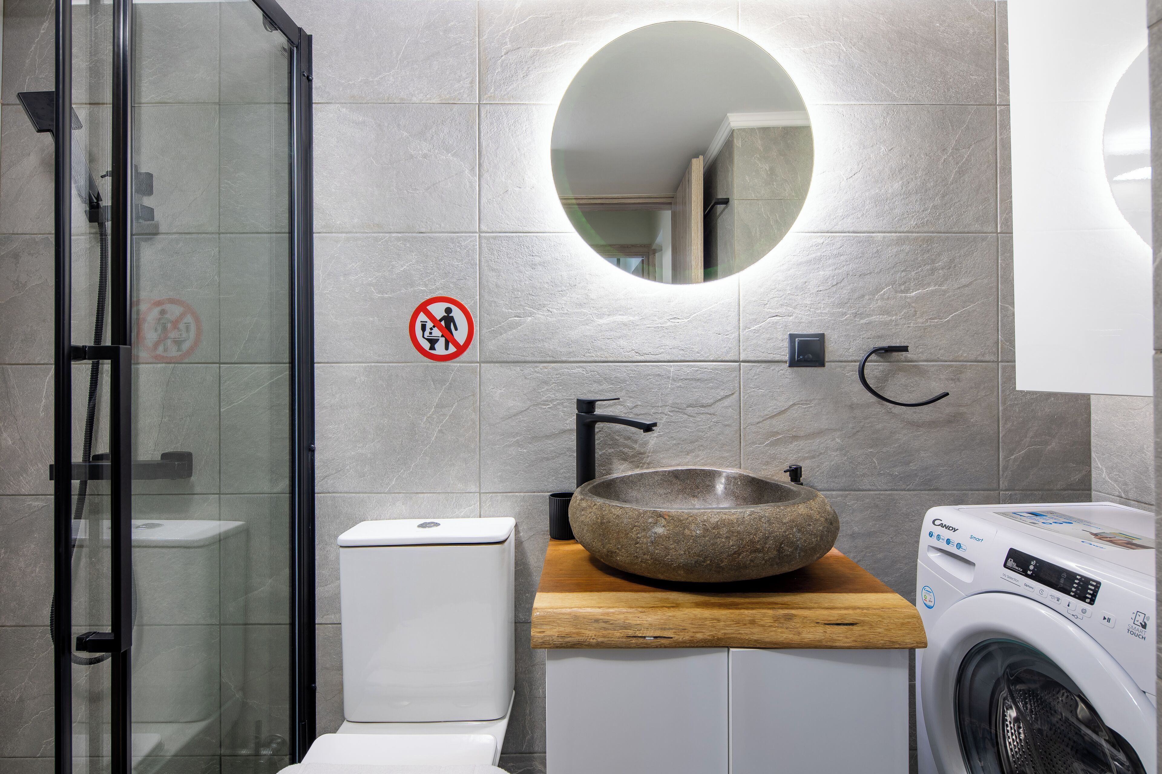 A modern bathroom with a glass-enclosed shower, a round mirror, a sink with a stone basin, a toilet, and a washing machine.